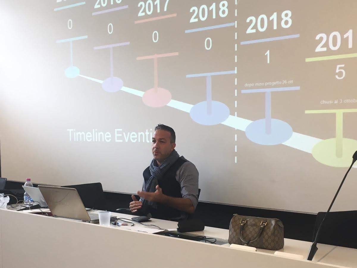 Relatore con sciarpa grigia alla cattedra con laptop, dietro di lui la slide proiettata «Timeline Eventi» con anni 2017-2018